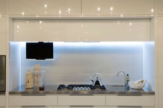 Compact contemporary kitchen with glossy white overhead and base cabinetry, a stainless-steel benchtop, and a pale illuminated splashback. The cooktop, kettle, sink, and wall-mounted television are integrated into the streamlined cabinetry.