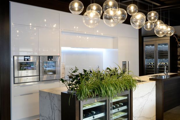 A sophisticated contemporary kitchen with glossy white full-height cabinetry, integrated stainless-steel appliances, a large marble-look island and a dark timber or black breakfast-bar section. A glass-fronted refrigerated wine or beverage cabinet sits beneath a central planter filled with greenery. Multiple spherical pendant lights hang from a dark ceiling, while additional tall refrigeration units are visible at the rear.