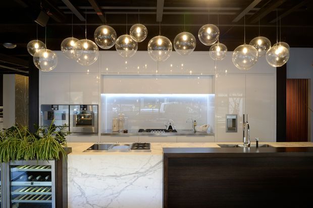 A sophisticated contemporary kitchen with a large marble-look island, dark timber breakfast-bar cabinetry, integrated appliances, and a backlit pale splashback. Numerous spherical glass pendant lights hang from an exposed dark ceiling grid, while a wine fridge and indoor greenery add visual interest.