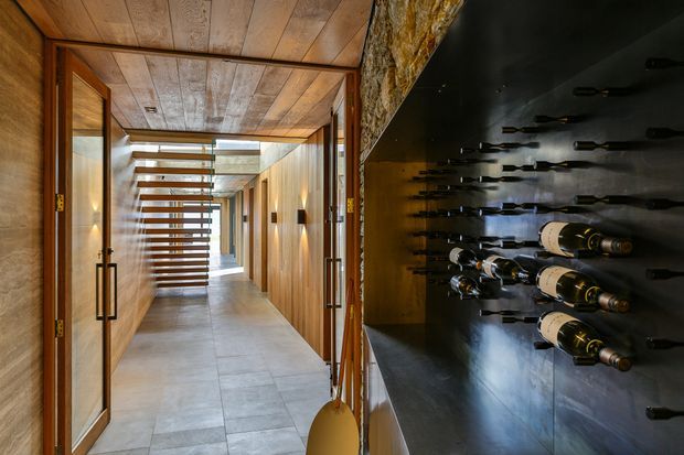 A warm, sophisticated interior corridor featuring timber wall and ceiling panelling, large grey stone-look floor tiles, illuminated wall sconces, and a prominent built-in black wine bottle display along the right wall. A slatted timber screen leads toward a brighter glazed area at the end of the passage.