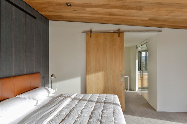 A bright contemporary bedroom with a partially visible bed, warm timber-lined ceiling, dark vertical wall panelling, and a simple orange-brown upholstered headboard. A large timber barn-style sliding door leads to a carpeted hallway and a sunlit adjoining area. The room has clean white walls, recessed ceiling lighting, and a warm, refined material palette.