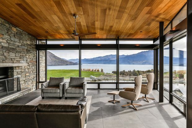 A contemporary living room with full-height glazing overlooking a lake and mountains. The space features a warm timber-clad ceiling, exposed stacked-stone fireplace wall, large-format grey floor tiles, black-framed glazing, a leather sectional sofa, sculptural lounge chairs, and a ceiling fan.