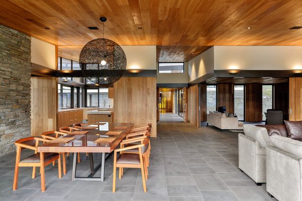 Open-plan interior featuring a dining area and living room with extensive warm timber wall and ceiling lining, a stacked-stone feature wall, dark timber-framed glazing, grey tiled flooring, and a large spherical pendant light. The space has a refined contemporary lodge-like character with strong connections to the outdoors through large windows and doors.