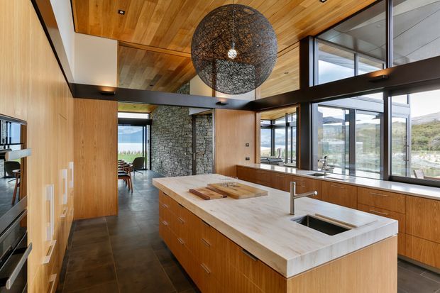 A spacious contemporary kitchen with warm timber wall and ceiling lining, dark stone or tile flooring, and extensive black-framed glazing. A large island with a pale stone benchtop, timber cabinetry, integrated sink and modern tap dominates the foreground. The kitchen opens toward a dining area and a rugged natural-stone feature wall, with landscape visible through the windows. A large woven spherical pendant light adds a sculptural focal point.