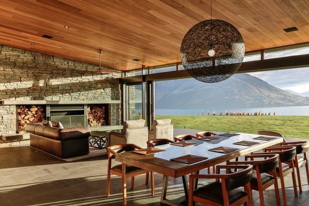Open-plan dining and living space with a large timber dining table, leather seating, stone fireplace wall with integrated firewood storage, timber-lined ceiling, extensive glazing, and a sculptural woven pendant light. A mountain and lake landscape is visible beyond the windows.