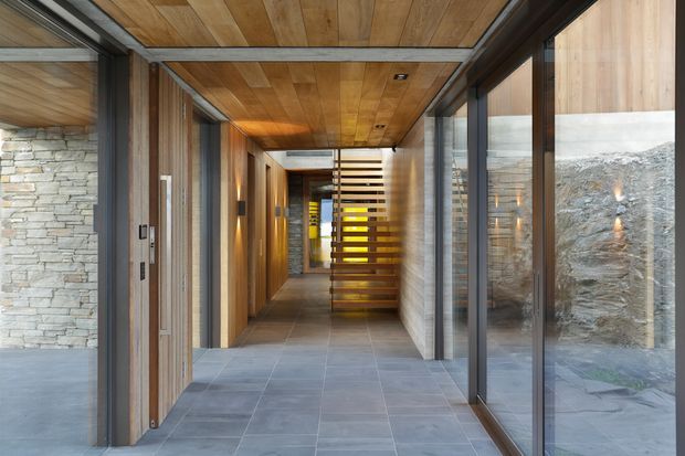 A modern interior hallway with a long grey tiled floor, timber-clad ceiling and walls, exposed stacked-stone feature walls, dark metal-framed glazing, and a sculptural open-riser timber stair at the far end. The corridor is well lit by concealed and recessed lighting, with views into adjacent spaces through full-height glass.