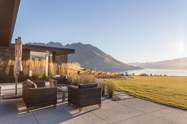 A contemporary lakeside outdoor terrace overlooking a mountain-backed lake. The paved patio has wicker lounge chairs, a low table, an umbrella, ornamental grasses, and a simple flat-roofed structure with extensive glazing and stone walls. Warm low-angle sunlight creates a calm, natural outdoor setting.