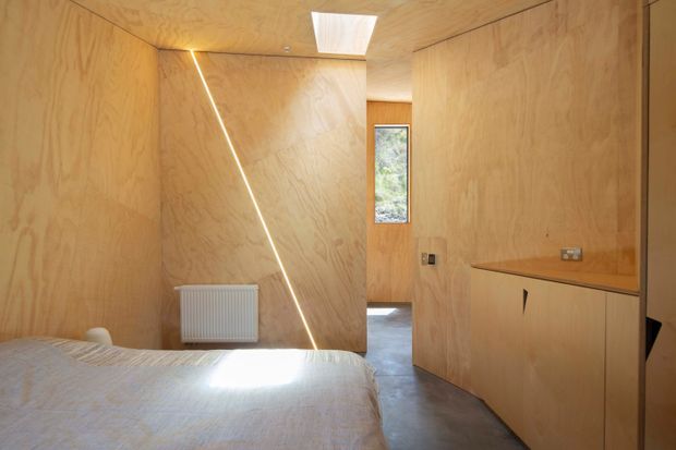 Compact plywood-lined bedroom with warm timber walls and ceiling, a partially visible bed in the foreground, built-in timber cabinetry along the right wall, polished concrete flooring, a narrow window, skylight, and a distinctive diagonal linear light or slot detail on the partition wall. The room has a restrained, highly integrated contemporary interior with natural daylight.