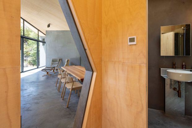 Interior view through a distinctive angled timber-lined opening, showing a dining area with a long wooden table and chairs, lounge chairs, full-height glazing, concrete flooring, and a compact dark-toned bathroom with a wall-mounted basin and mirror.