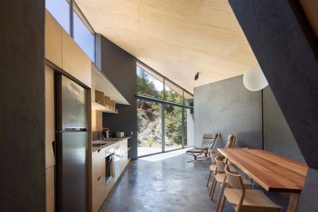 A bright, open-plan modern kitchen and dining area with a polished concrete floor, timber cabinetry and ceiling, dark painted walls, expansive floor-to-ceiling glazing, and a long timber dining table. The space includes a stainless-steel refrigerator, kitchen island, sculptural pendant light, and mid-century-inspired dining chairs and lounge chairs. Natural rocky terrain and vegetation are visible through the glazing.