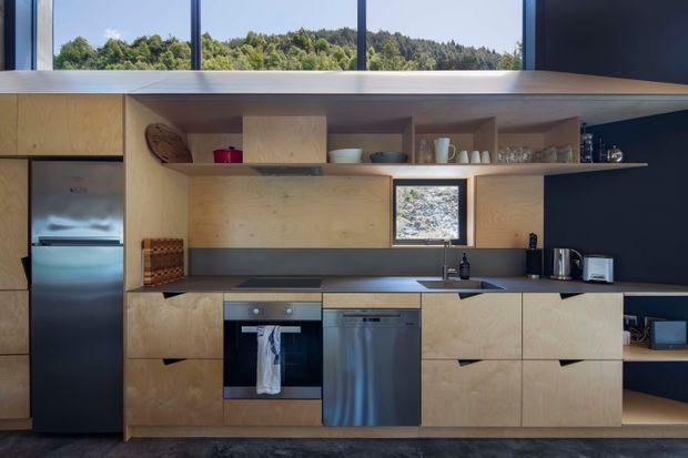 A compact contemporary kitchen with light plywood cabinetry, integrated shelving, dark charcoal splashback and wall surfaces, stainless-steel refrigerator and dishwasher, built-in oven, induction cooktop, and a long central worktop with sink. A high horizontal window brings in daylight and overlooks wooded hills, while open shelves display crockery and glassware.
