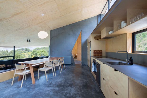 Open-plan kitchen and dining area with plywood-lined ceiling and cabinetry, dark concrete flooring, a long timber dining table, and expansive glazing overlooking greenery. The kitchen features a dark benchtop, integrated sink, open timber shelving, and a sculptural timber doorway or partition against a dark feature wall.