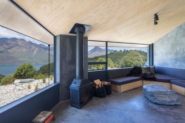 A contemporary panoramic living room with floor-to-ceiling glazing overlooking a mountain lake and forested landscape. The room has a built-in L-shaped upholstered bench, a freestanding black wood-burning stove with flue, a pale timber ceiling, polished grey concrete flooring, dark window frames, and a minimalist low-profile lounge arrangement.