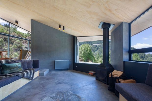 A contemporary living room with expansive black-framed glazing overlooking a wooded landscape. The room features polished concrete flooring, exposed plywood ceiling panels, dark concrete feature walls, a freestanding wood-burning stove with flue, and understated upholstered seating.