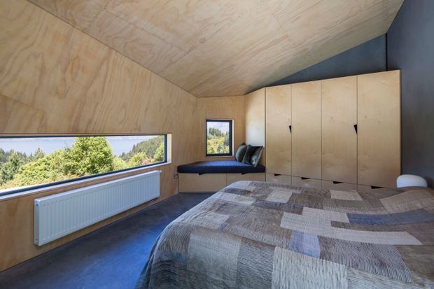 Compact modern bedroom with plywood-lined walls and ceiling, a large horizontal corner window overlooking greenery, built-in timber cabinetry, a window-side bench seat, dark flooring, and a patchwork bedspread. The room has a warm natural-material palette with strong geometric built-in elements and abundant daylight.