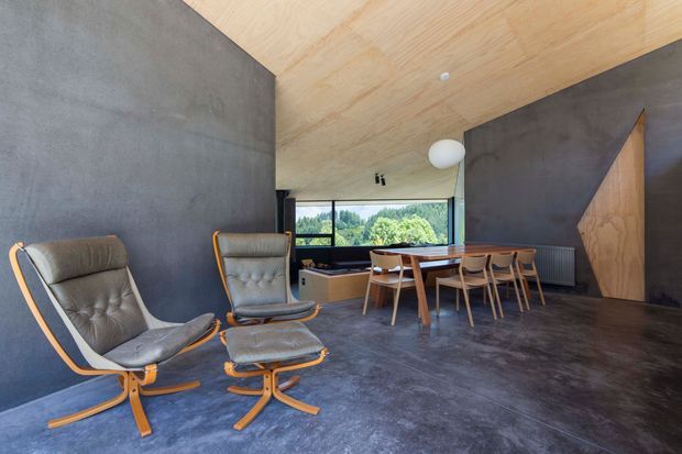 A spacious contemporary dining and living area with dark charcoal concrete walls and flooring, a warm plywood ceiling, and a large horizontal window overlooking greenery. Two leather lounge chairs with timber frames sit beside a long timber dining table surrounded by matching chairs. A sculptural white pendant light, exposed timber joinery, and a radiator complete the restrained, material-focused interior.