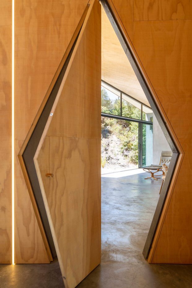 A dramatic geometric plywood pivot door creates a faceted opening between a warm timber-lined interior and a bright concrete-floored living space. Large glazed openings bring in natural light and reveal a rocky landscaped exterior, while a sculptural lounge chair is visible beyond.