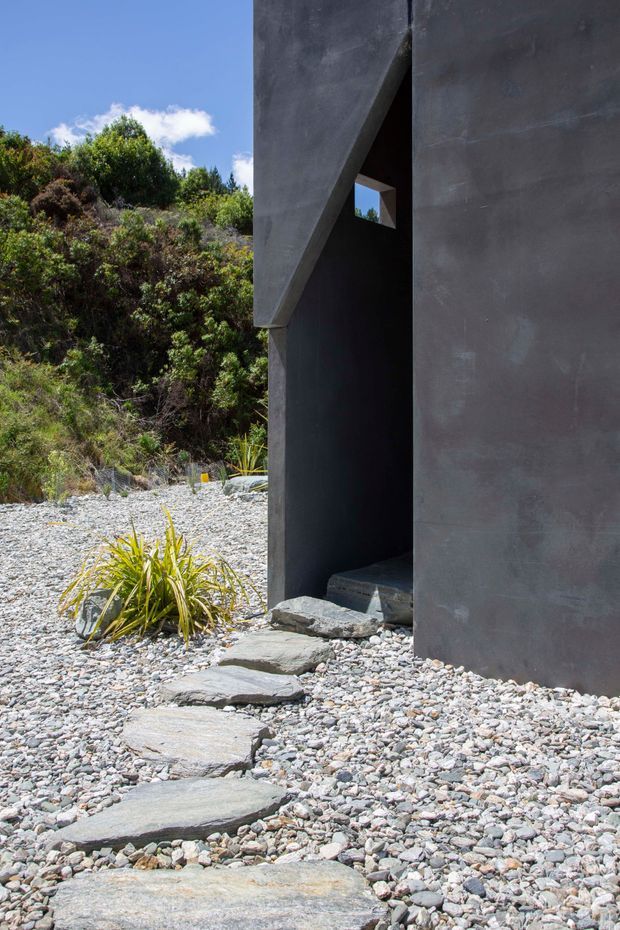 Exterior view of a stark contemporary structure with dark charcoal concrete or plastered walls, a deeply recessed angular entry, and a small square opening. Large irregular stepping stones cross a pale grey gravel landscape with hardy native-style planting and a vegetated hillside beyond. The composition emphasizes monolithic geometry, shadow, and a naturalistic minimalist setting.