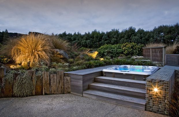 A landscaped outdoor spa area set into a naturalistic garden, with a raised hot tub surrounded by timber decking, broad timber steps, gabion-style stone seating or retaining elements, ornamental grasses, shrubs, and low garden lighting. The scene has a rustic, warm, and tranquil atmosphere under an overcast evening sky.