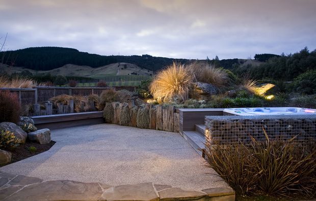 A landscaped outdoor terrace or courtyard at dusk, featuring a broad paved entertaining area, built-in timber seating and raised planters, ornamental grasses and sculptural planting, natural stone edging, and a raised spa or hot tub enclosed in a gabion-style metal frame. Rolling wooded hills form the distant backdrop.