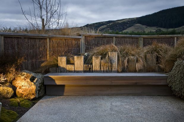 A landscaped outdoor terrace or courtyard with a long built-in timber bench, weathered timber privacy fencing, natural stone edging, gravel paving, and ornamental grasses. The setting overlooks a rugged, forested mountain landscape beneath an overcast sky, creating a natural, sheltered outdoor seating area.