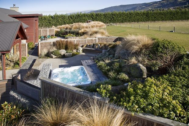 A landscaped outdoor courtyard beside a modern timber-clad house, featuring a small raised spa pool, layered native-style ornamental grasses, shrubs, stone-edged planting beds, and timber privacy fencing. The property sits beside open fields with a mountainous backdrop.