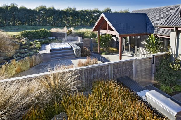 Elevated exterior view of a landscaped residential courtyard and entertaining area, featuring a timber-clad building with steep gabled metal roofing, a covered entry or outdoor pavilion, a raised spa or plunge pool, timber screening fences, and dense ornamental grasses and sculptural plantings.