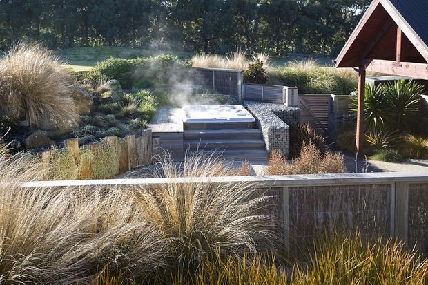 A landscaped outdoor spa area with a steaming hot tub set into a raised timber-deck platform and reached by broad steps. The space is screened by timber fencing and surrounded by ornamental grasses, native-style planting, and a small timber gable-roof structure at the right.