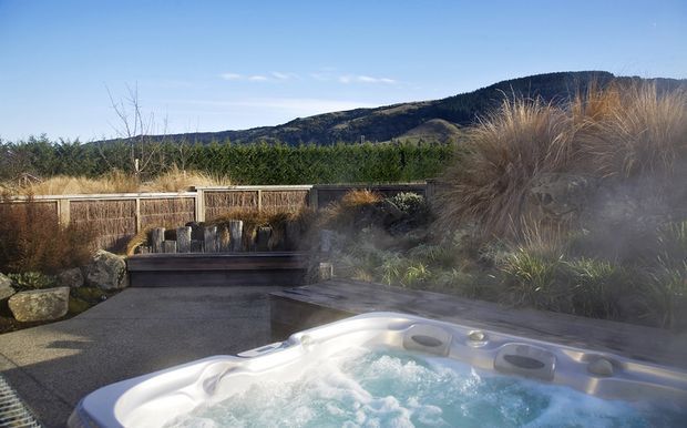An outdoor spa pool bubbles in the foreground of a landscaped garden, with ornamental grasses, low stonework, timber screening, and a distant forested hillside under a clear blue sky.