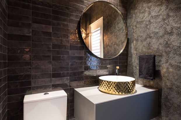 A compact, dark-toned bathroom featuring glossy charcoal rectangular wall tiles, a textured grey feature wall, a round mirror with a slim metallic frame, a white toilet, and a floating vanity with a white countertop and distinctive gold-and-black patterned vessel basin. The space has a dramatic, refined contemporary feel with warm reflected light.