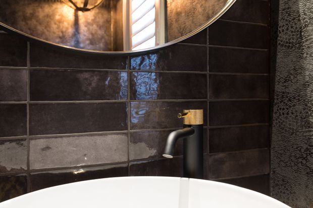 Close-up of a dramatic bathroom vanity area featuring a white basin, matte black wall-mounted tapware, glossy dark rectangular wall tiles, and a round mirror. A textured dark wall surface and warm reflected lighting add contrast and depth.