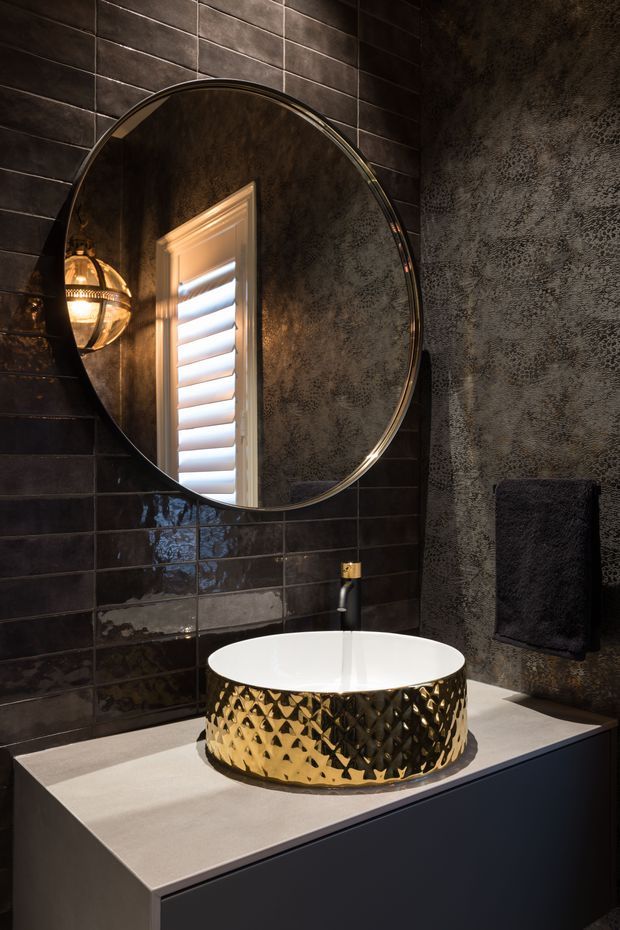 A moody, luxurious bathroom vanity with a round black-framed mirror, dark glossy rectangular wall tiles, textured dark feature wall, and a sculptural gold hammered vessel basin on a pale stone benchtop. A warm wall sconce and reflected shuttered window add atmosphere, with dark integrated cabinetry below.