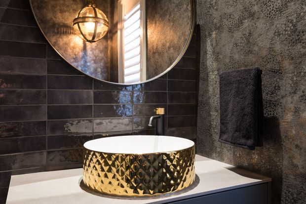 A dramatic, luxurious bathroom vanity featuring a freestanding round vessel basin with a textured metallic gold finish, set on a pale stone benchtop. Dark glossy rectangular wall tiles, a heavily textured dark feature wall, a black wall-mounted tap, a large round mirror and a spherical pendant light create a moody, high-contrast interior.