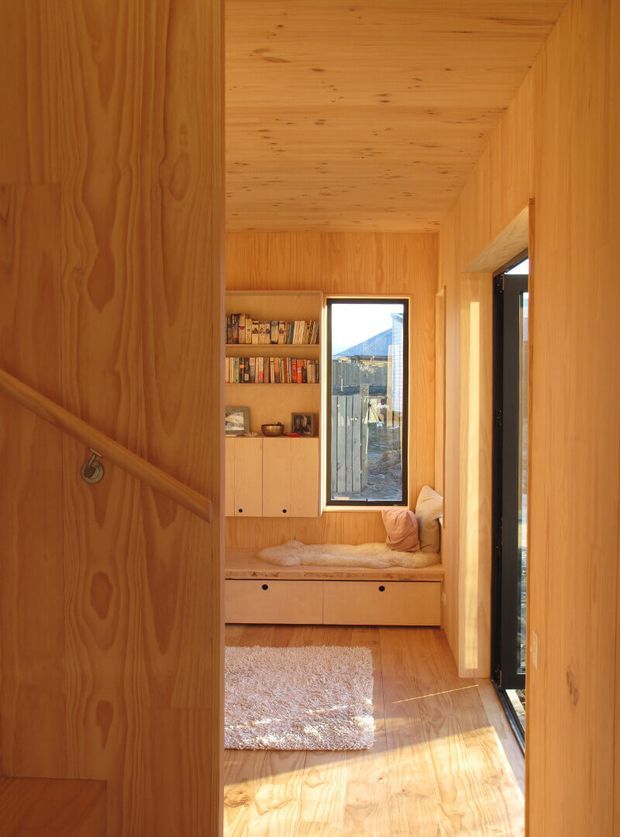 Warm, compact timber-lined interior hallway or entry nook with built-in cabinetry, open bookshelves, a window seat with cushions and drawers below, and a pale textured rug. Vertical timber wall and ceiling panelling create a cohesive natural-wood enclosure, while a glazed door or opening on the right brings in strong sunlight and views outside.