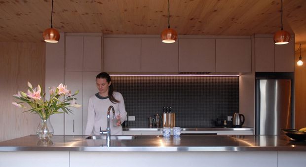 A contemporary kitchen viewed across a broad island bench, with pale full-height cabinetry, a dark small-format tiled splashback, stainless-steel appliances, timber-lined ceiling and walls, and three copper pendant lights. A woman stands behind the island near the sink, with flowers and small kitchen items on the benchtop.