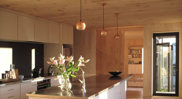 Warm, timber-lined contemporary kitchen with a large central island, dark benchtop, pale fixed cabinetry, integrated appliances, pendant lighting, and a glazed exterior door. A timber-lined passage or living area is visible beyond the kitchen.