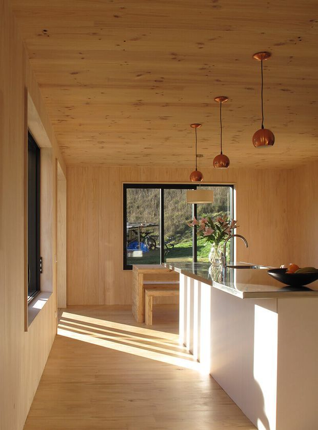 A warm, light-filled contemporary kitchen with extensive natural timber lining on the walls and ceiling, timber flooring, and minimalist built-in cabinetry. A large island with a pale benchtop occupies the foreground, paired with a black sink and curved tap. Three copper-toned pendant lights hang above the island, while black-framed glazing opens views to greenery outside. Strong afternoon sunlight creates long geometric shadows across the floor and cabinetry.