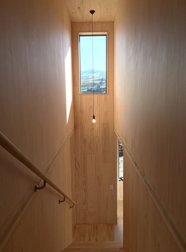 A narrow timber-lined stairway descending between warm wood-panelled walls. A simple pendant light hangs in front of a tall window framing a mountainous rural landscape, while a timber handrail follows the left wall. The space has a restrained, natural, contemporary feel with strong vertical lines and warm daylight.