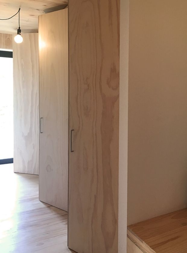 A warm, minimalist interior detail showing full-height light timber built-in cabinetry with simple dark handles. The cabinetry forms a partition or storage wall beside an open doorway, with matching timber flooring and ceiling, a plain warm neutral wall, and a simple exposed pendant light. Daylight enters from the left.