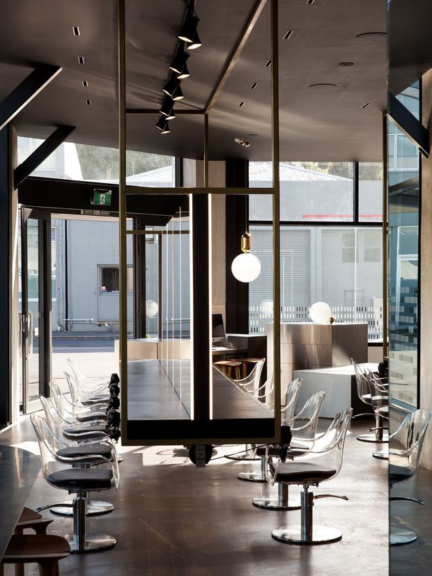 A sophisticated commercial interior with rows of transparent acrylic chairs arranged around long tables, framed by dark timber and bronze-toned glazed partitions. The space has polished concrete flooring, a dark ceiling with recessed lighting and black track pendants, sculptural globe lights, and extensive glazing overlooking an adjoining courtyard or building. The composition suggests a contemporary café, restaurant, or hospitality venue.