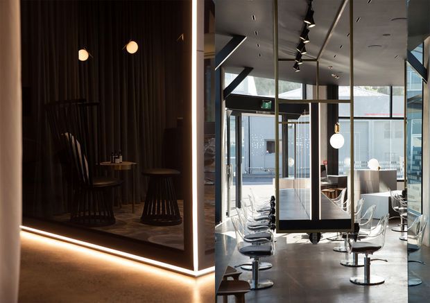 A contemporary commercial interior, likely a café, hospitality venue, or showroom, featuring a dramatic dark timber-lined alcove with small round tables and stools beside a bright glazed seating area. The space includes exposed structural framing, floor-to-ceiling glazing, polished concrete flooring, suspended track lighting, pendant lights, and modern transparent chairs and bar stools.