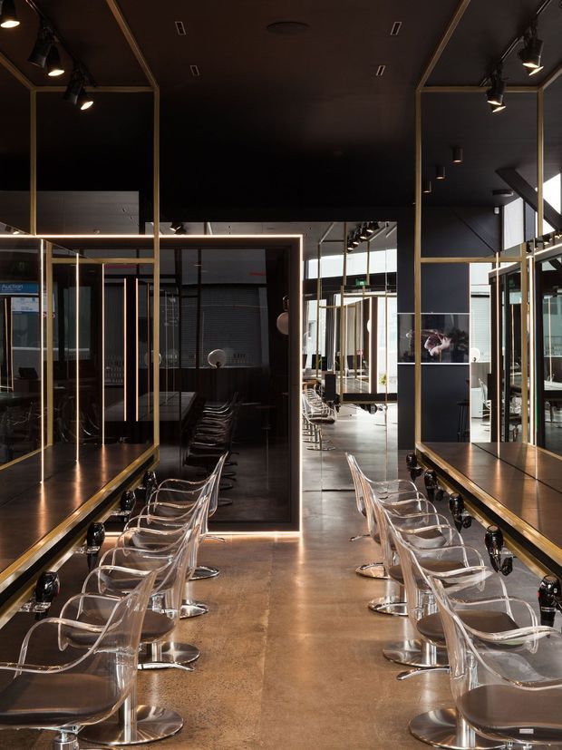 A dark, dramatic contemporary commercial salon interior with two rows of transparent salon chairs facing long mirrored styling stations. The space features black walls and ceilings, warm metallic gold-framed mirrors and partitions, integrated strip lighting, polished concrete flooring, and exposed structural elements with abundant reflections.