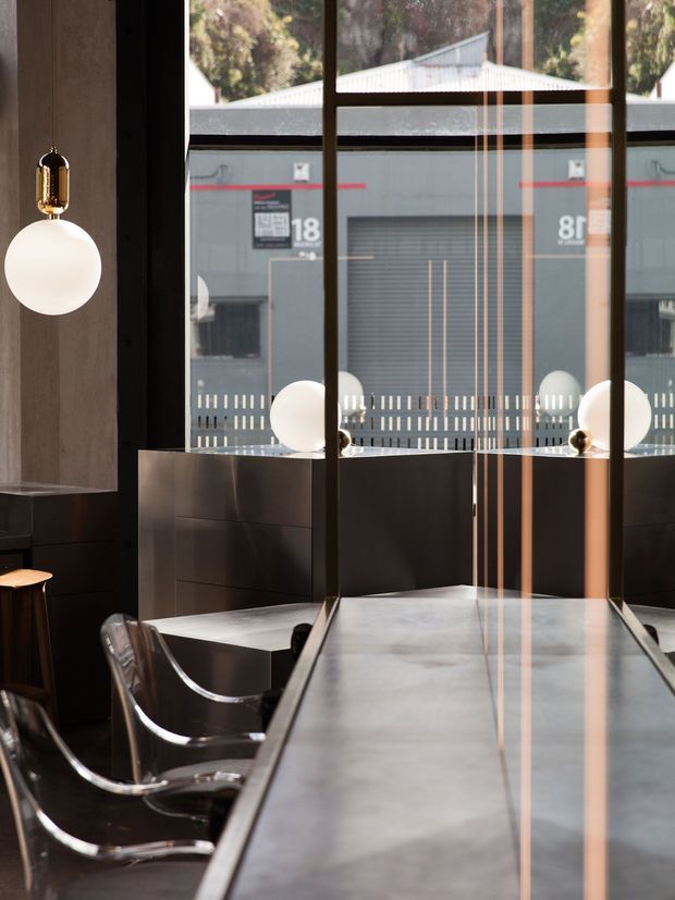 A refined modern dining or hospitality interior with a long dark stone or metal table, transparent sculptural dining chairs, dark built-in cabinetry, globe pendant lights, and full-height black-framed glazing. The glazing reflects the interior and reveals an industrial-looking building outside.