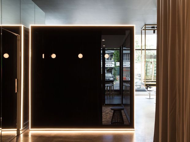A dramatic contemporary interior featuring a large black reflective partition or framed wall with integrated warm LED edge lighting. Pendant lights hang within the dark zone, while a brighter open area with glazing, seating, and architectural finishes is visible beyond. Floor-to-ceiling curtains frame the right side, and the polished floor and dark finishes create a moody, high-contrast atmosphere.
