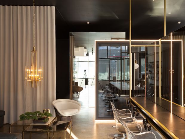 A sophisticated, dark-toned salon or commercial interior with a long mirrored styling counter, transparent chairs, warm integrated lighting, and a dramatic gold-framed partition. A sculptural chandelier hangs beside full-height cream curtains, while polished concrete flooring and reflective surfaces create a refined, high-contrast atmosphere.