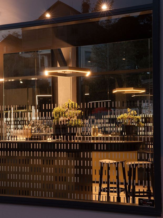A dimly lit, sophisticated bar or hospitality interior viewed through large glazing. The space features reflective glass surfaces, dark timber or metal screens with repeated vertical slots, warm linear pendant lighting, a counter with bottles and glassware, decorative potted plants, and high stools. Reflections reveal additional seating and architectural elements, creating a layered, intimate atmosphere.
