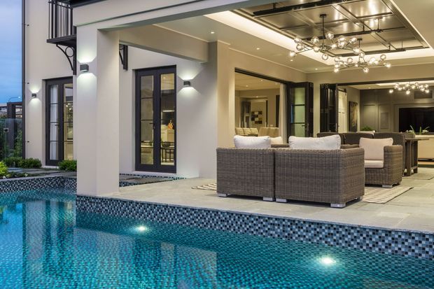 A contemporary outdoor entertaining area beside a tiled swimming pool, with a covered lounge and dining space opening through large glazed doors to the pool. The architecture features clean white rendered walls, dark-framed glazing, a flat roof, and a striking exposed dark ceiling with decorative pendant lighting. Wicker-style outdoor sofas and chairs create a refined resort-like setting.