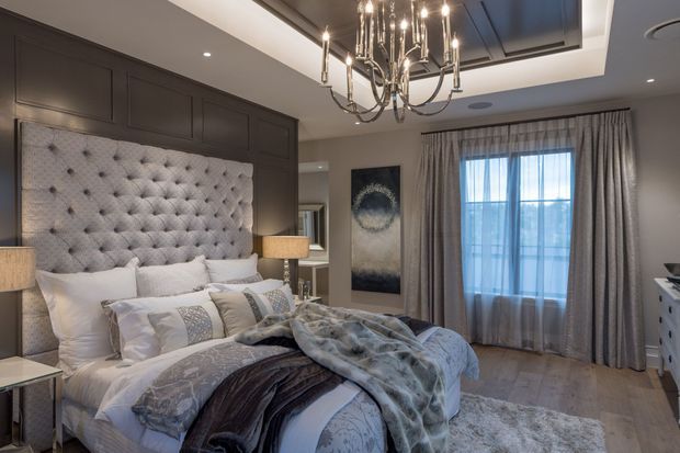 A refined bedroom with a large upholstered tufted headboard integrated into dark built-in wall panelling. The bed is dressed in layered white, grey, and taupe bedding, with bedside lighting, a dramatic multi-arm chandelier, full-height curtains, pale timber flooring, and a soft area rug. The room has a polished, luxurious and slightly moody atmosphere.