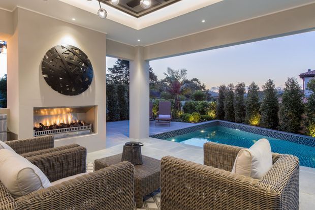 A refined covered outdoor lounge overlooking a swimming pool and landscaped garden. The space features woven rattan-style lounge chairs and a low coffee table, a built-in linear fireplace with a circular sculptural wall feature, recessed lighting, and broad open glazing or openings to the garden. The palette is neutral and warm, with a relaxed resort-like atmosphere.