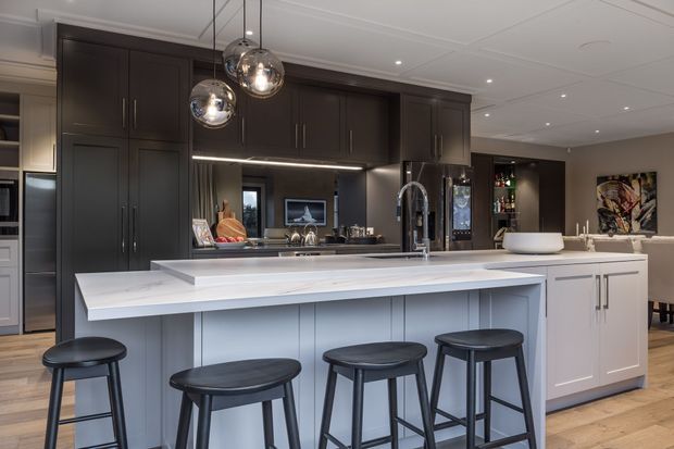 A sophisticated open-plan kitchen dominated by a large white island with integrated sink and seating for four, paired with dark timber-toned full-height cabinetry and a mirrored splashback. Pendant globe lights hang above the island, with a dining and living area visible beyond. The room has warm timber flooring, recessed ceiling lighting, and a refined contemporary fit-out.
